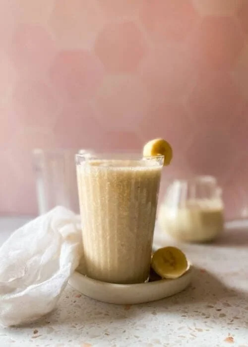 Easy Korean Banana Milk Recipe - Super Creamy 바나나 우유 5 close up banana milk in a glass, on a tray with sliced banana, a towel, another glass and jug behind, a pink and speckled background