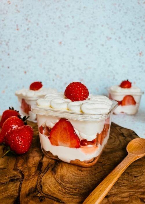 mini strawberry cake pots with strawberries around, a spoon, on a wooden board, speckled background