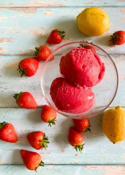 from top strawberry sorbet no churn in a glass bowl, strawberries and lemons around on a light blue background
