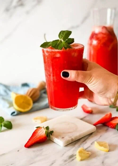 Easy Blended Strawberry Lemonade Recipe 2 hand holding the. glass of blended strawberry lemonade on a marble coaster, behind a jug of lemonade, strawberries mint and lemons around, in the back a blue towel and lemon squeezer on a marble background