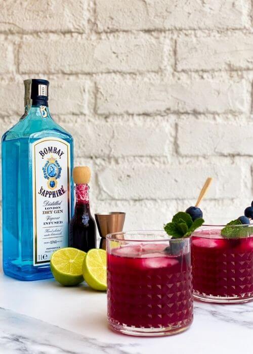 Amazing Blueberry Gin Cocktail, A 10-Minute Tasty Gin Tonic 2 two glasses of blueberry gin cocktail, in the back lime, blueberry syrup and measure cup, on a white and marble background, bottle of sapphire gin