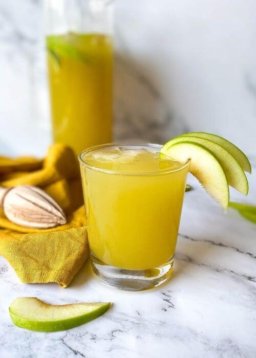 Refreshing Green Apple Mocktail, 3 Ingredients 3 close up on a marble background, a glass of green apple mocktail, beside a yellow towel and lemon squeezer, behind a jug of green apple mocktail, slices of apples around