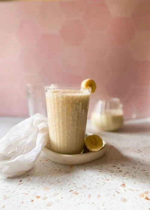 Easy Korean Banana Milk Recipe - Super Creamy 바나나 우유 4 banana milk in a glass, on a tray with sliced banana, a towel, another glass and jug behind, a pink and speckled background