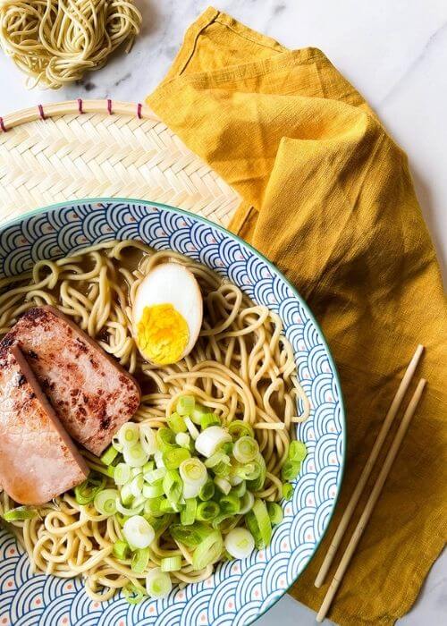 a bowl of ponyo ramen with noodles, spring onions, ham and egg. under a yellow towel, chopsticks, bowls with ingredients on a marble background