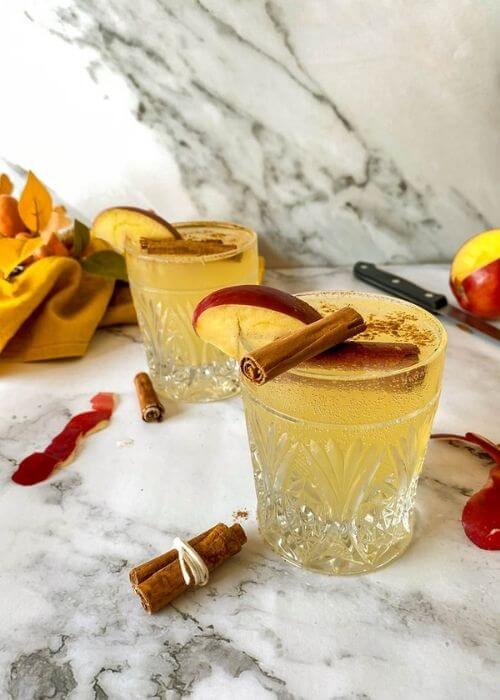 Quick Apple Fizz Mocktail, Delicious Fall Drink 1 apple fizz mocktail with apple slices and cinnamon sticks on top, apple peels, cinnamon sticks, knife, towel and leaves around, on marble background, glass bottle of ginger beer