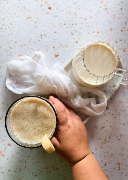 Easy Korean Banana Milk Recipe - Super Creamy 바나나 우유 3 from top, hand reaching for a glass of banana milk, towel and jug of milk beside, on a marble tray, on speckled background