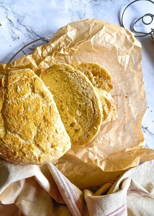 no knead bread, under parchment paper, wire rack, towel and on the side a bread a dough whisk, on marble background, slices pn the paper