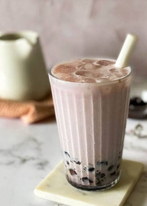 taro latte in a glass with a straw, on a marble coaster, jug, towel and boba pearls in the background, on a marble background