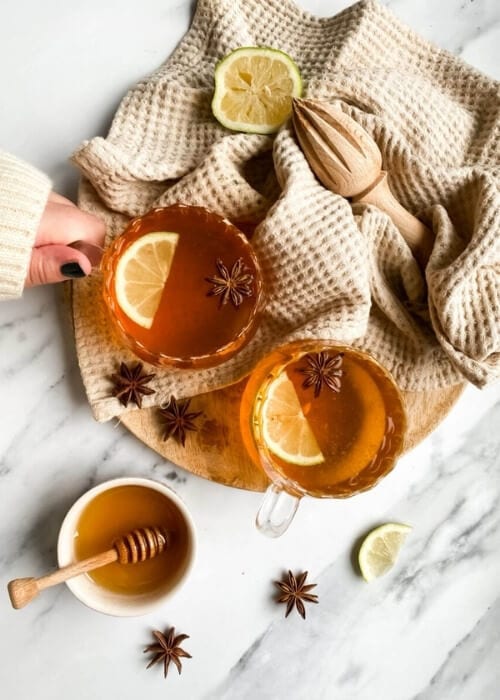 Festive Hot Toddy Mocktail Recipe, Warm & Delicious 1 from top, two cups of hot toddy mocktail with slices of lemon and star anise, over a wooden board and light brown tea towel, slices of lemons, star anise and honey around, hand holding one cup,