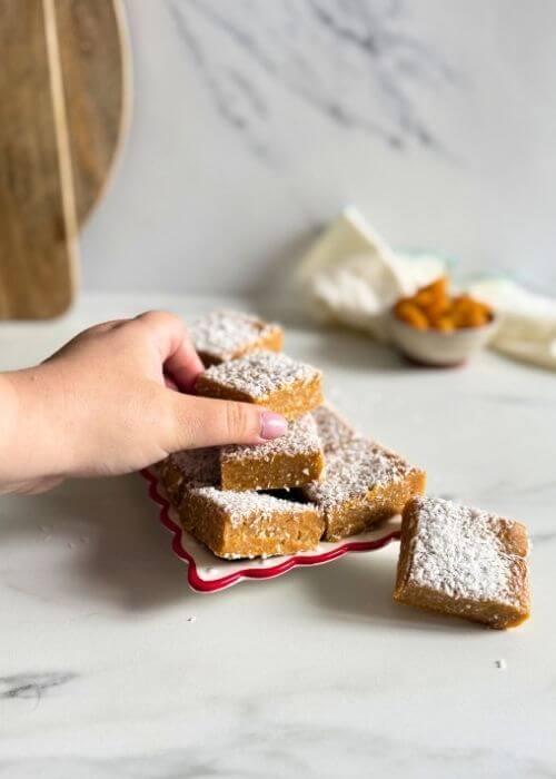 hand holding a slice of apricot slice, on a plate more apricot slice, behind a bowl of apricots, towel and two wooden boards on a marble background