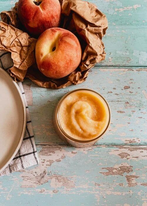 from top peach curd in a jar on a plate, under a towel, in the back two peaches on a paper bag