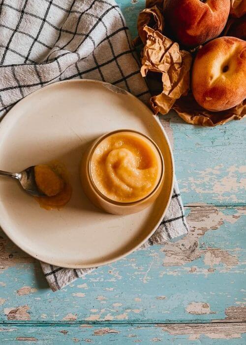 from top peach curd in a jar on a plate with a spoon, under a towel, in the back two peaches on a paper bag