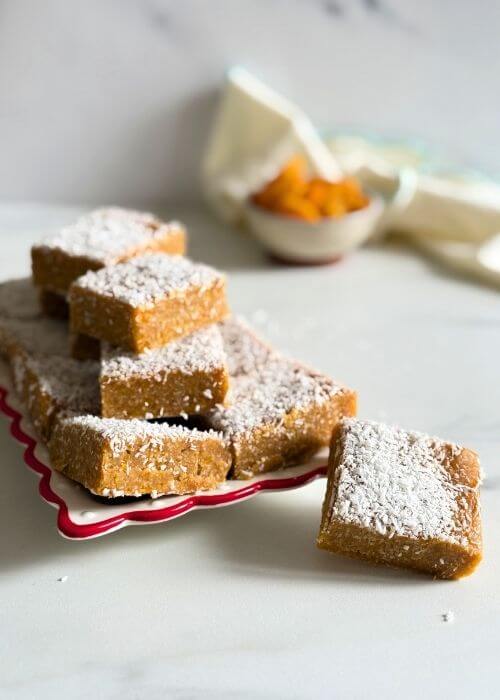 close up on a plate more apricot slice, behind a bowl of apricots, towel and two wooden boards on a marble background