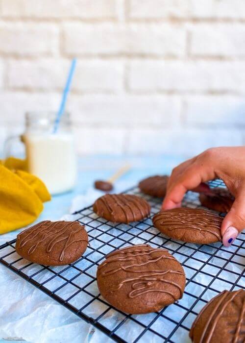 hand reaching ifor 3 ingredient nutella cookies on a cookie rack, a spoon with nutella, glass of milk, yellow napkin