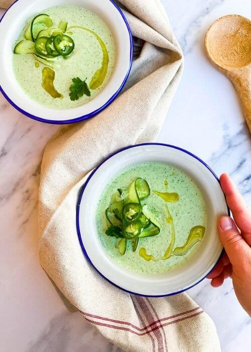 from top - two bowls of cucumber gazpacho on a towel, spoon on the side, on marble background, hands holding one bowl