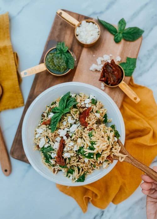 Ultimate Easy Orzo Pesto Salad Recipe For A Delicious Summer 2 hand holding a spoon of orzo salad in a bowl with a spoon, pesto, sundried tomatoes and feta in measuring cups, on a wooden board, around a yellow towel