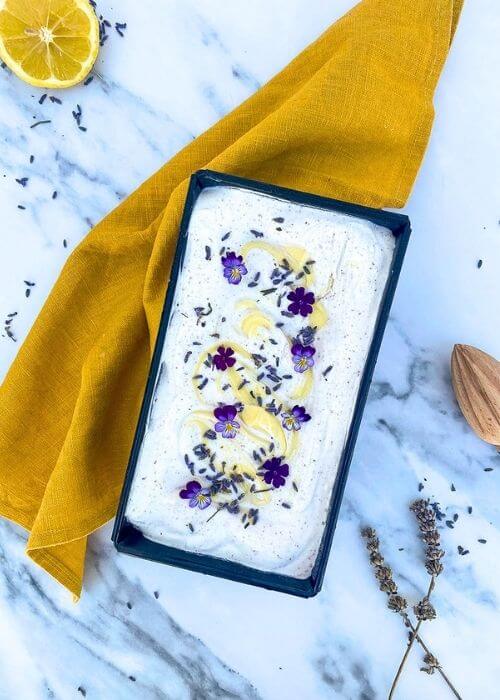 from top - lavender lemon ice cream in a loaf pan, a yellow towel under, lemon, lemon squeezer and lavender buds around, on a marble background