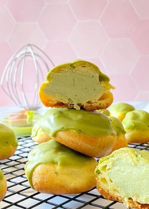 close up of a stack of matcha cream puffs on a wire rack, behind a whisk, a white towel on a pink tiled background