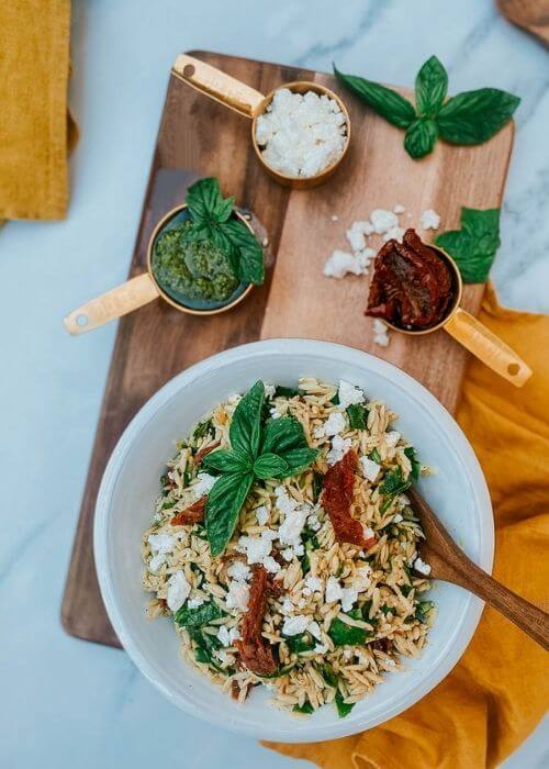 Ultimate Easy Orzo Pesto Salad Recipe For A Delicious Summer 4 orzo pesto salad in a bowl with a spoon, pesto, sundried tomatoes and feta in measuring cups, on a wooden board, around a yellow towel