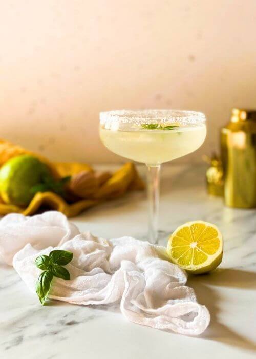 Easy Basil Lemon Martini - Delicious 10 Minutes Vodka Drink 3 from front basil lemon martini in a glass on a marble background, on the side a lemon, some basil leaves and a white towel, in the background a yellow towel, lemon squeezer and cocktail shaker