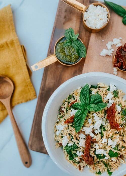 Ultimate Easy Orzo Pesto Salad Recipe For A Delicious Summer 3 close up of orzo salad - pesto, sundried tomatoes and feta in measuring cups, on a wooden board, around a yellow towel