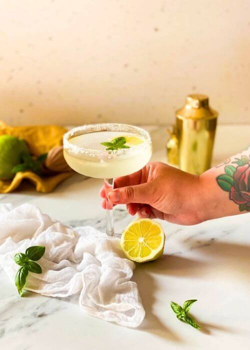Easy Basil Lemon Martini - Delicious 10 Minutes Vodka Drink 5 hand holding a basil lemon martini in a glass on a marble background, on the side a lemon, some basil leaves and a white towel, in the background a yellow towel, lemon squeezer and cocktail shaker