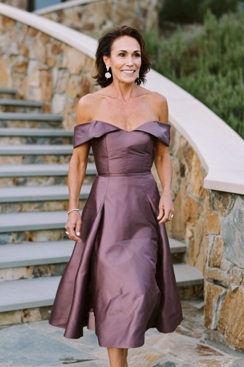 A woman over 50 wears a mauve off-the-shoulder formal dress with a structured bodice and soft flare skirt, styled with vintage drop earrings and block heels