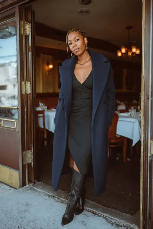 A Black woman over 40 wears a navy wool wrap coat over a sleek black midi dress, paired with tall leather boots and subtle silver jewelry 