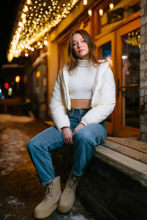A curvy woman wears a simple white ribbed turtleneck with a cropped puffer jacket, straight jeans, and warm winter boots, posing outside a cozy dinner café with string lights