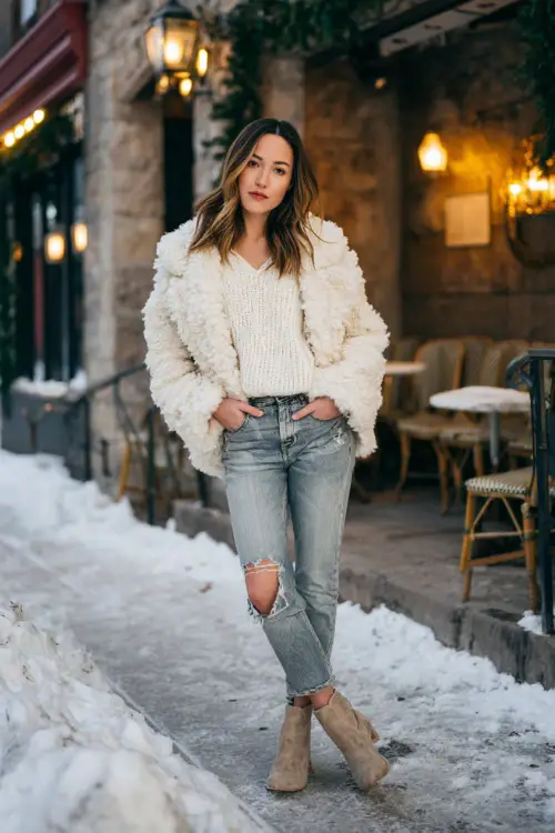 A woman wears a cozy white knit sweater with medium-wash jeans, suede ankle boots, and a faux-fur jacket, posing on a snowy sidewalk in front of a romantic dinner spot 