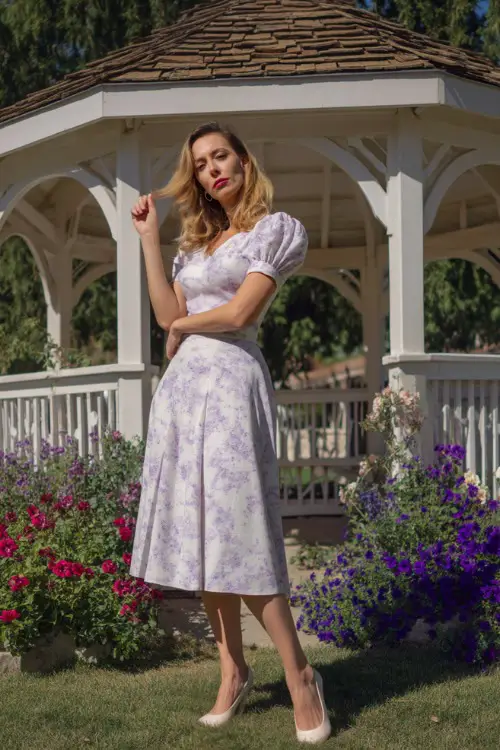 A poised woman over 50 in a white and lavender floral tea-length dress with puff sleeves and low heels, posing in front of a small white gazebo with spring flowers all around 