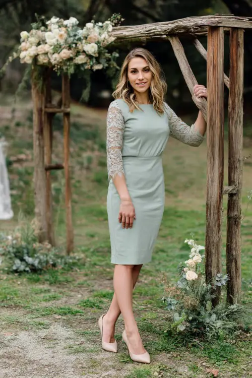 A woman over 50 in a soft sage green midi dress with lace sleeves and beige pumps, standing near a rustic wooden wedding arch with light spring florals