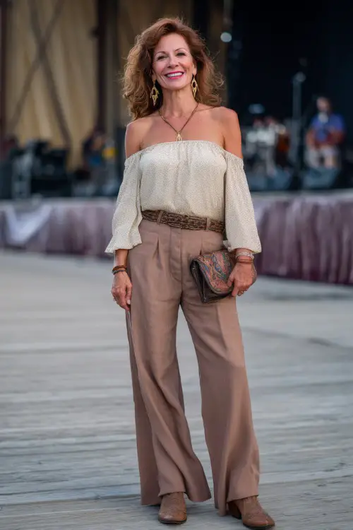 A woman over 40 styled in wide-leg linen pants, a fitted off-shoulder blouse, and tall embroidered cowboy boots, accessorized with boho earrings and a leather clutch 