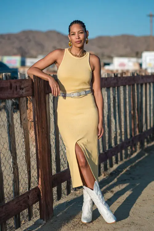 A woman over 40 wearing a simple ribbed tank midi dress with a slit, western belt, and white cowboy boots, with gold hoop earrings and a low bun 