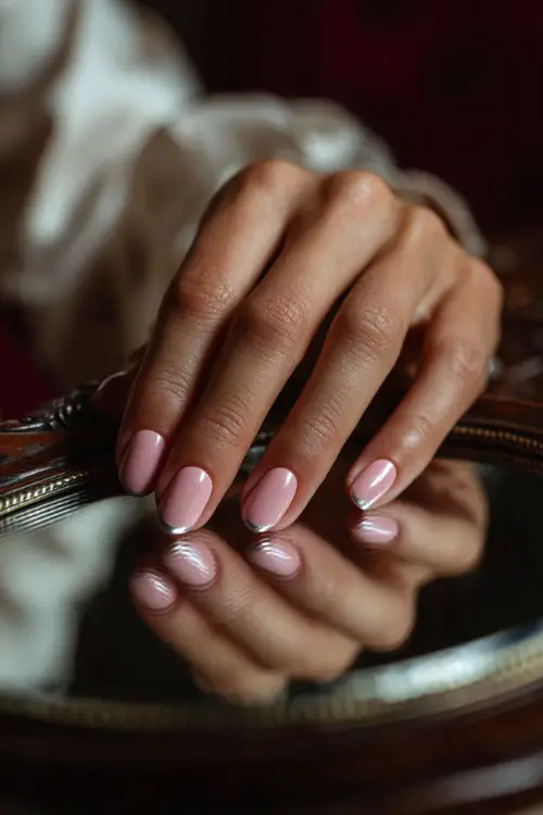 Bride’s hand brushing over mirror, short oval nails in glassy pastel pink with chrome shimmer tip on one nail, classic vanity background 
