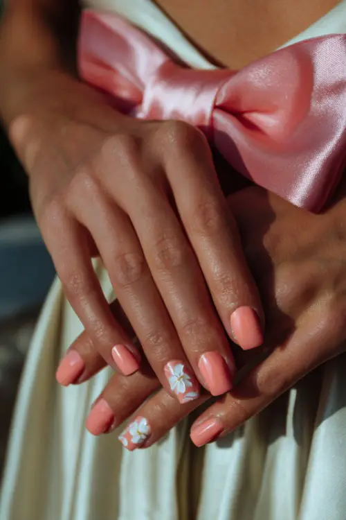 Bride’s hand holding satin bow on gown, short pastel coral nails with a glossy finish and one white floral decal