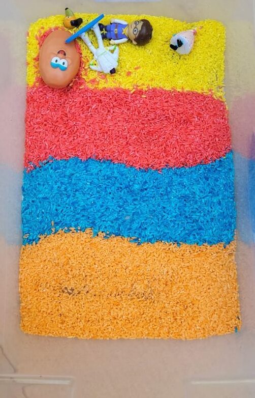 Yellow, Red, Blue and Orange rice in horizontal lines in bin with toys
