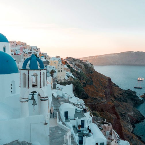 Eine malerische Ansicht von Oia, Santorini, mit den ikonischen weiß getünchten Gebäuden und ihren leuchtend blauen Kuppeln, die sich an die Klippen schmiegen. Im Hintergrund erstreckt sich das ruhige Meer mit einem Segelschiff und fernen Bergen unter einem klaren Himmel mit sanften Farbtönen.