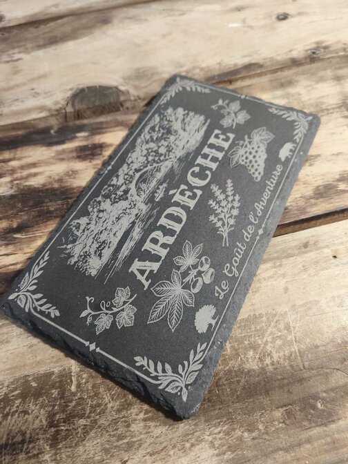 Ardoise rectangulaire gravée au laser par Gravlaz en Ardèche, reposant sur une table en bois brut. La gravure de couleur gris clair représente, en haut, le Pont d'Arc et les gorges, au centre, le texte "ARDÈCHE" en grandes lettres stylisées, et en bas, des motifs d'olivier, lavande et vigne avec l'inscription "Le Goût de l'Aventure".