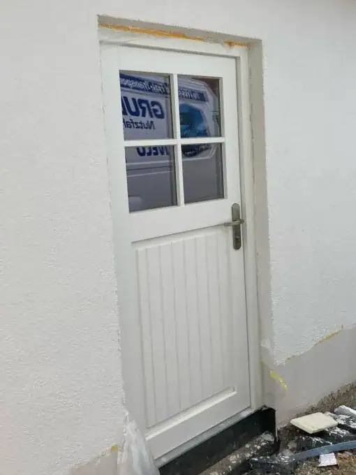 Weiße Nebeneingangstür von Fütterer Fenster & Türen GmbH mit vierfachen Glasausschnitt im oberen Bereich und vertikaler Paneeloptik im unteren Bereich, eingebaut in eine weiße Wand.