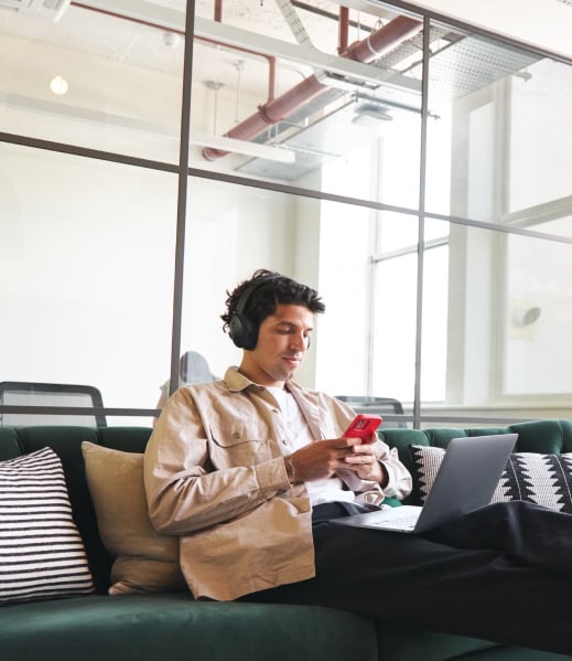 A man sitting on a sofa. He has his laptop on his lap and is looking at the phone in his hands.