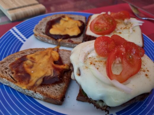 Burger bzw. Fleischkäse mit Schmelzkäse, Zwiebeln, Tomaten und nichts sichbar darunter Gurke auf Brot