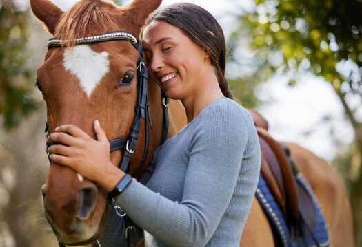 Woman affectionately embraces a brown horse, both smiling in a sunny outdoor setting.