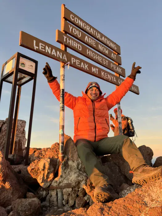 A man in hiking gear sits in front of a sign denoting the third highest peak of Mount Kenya.