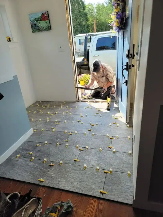 A handyman in Coeur D'Alene installing gray floor tiles with yellow leveling spacers in a home entryway with a truck visible in the background.