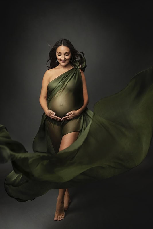 Elegant gravidfotografi av en smilende, gravid kvinne i grønt stoff, som omfavner sin mage i en studio setting, perfekt for elegante og minnerike gravidfotograferinger.
