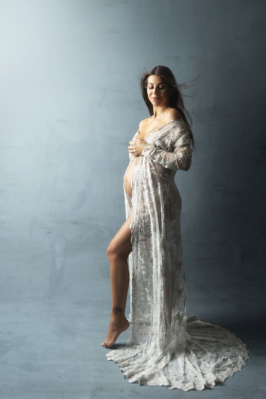 Elegante gravidfoto av kvinne i hvit stoffkjole som viser mage, mot en enkel blå bakgrunn, perfekt for gravidfotografering og minneverdige øyeblikk.