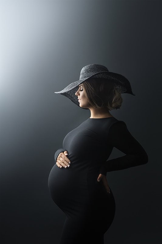 Elegant gravidfotografering av gravid kvinne i svart kjole med bredbremmet sommerhatt, kunstnerisk belysning og minimalistisk bakgrunn, perfekt for gravidfoto og mammatografi.