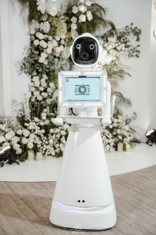 White robot photo booth standing in front of a floral wedding backdrop