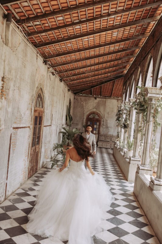 Bride running toward groom in cloister with chequered marble floor, Benedict Studio Bangkok